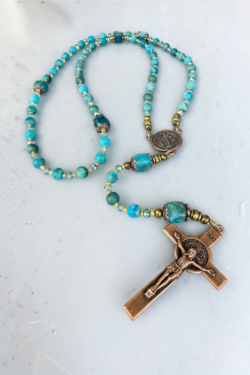 A beaded rosary necklace featuring turquoise-blue and light blue round beads, accented with small gold spacer beads and a bronze-finished crucifix pendant with a detailed figure of Jesus.