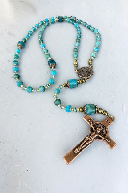 A beaded rosary necklace featuring turquoise-blue and light blue round beads, accented with small gold spacer beads and a bronze-finished crucifix pendant with a detailed figure of Jesus.